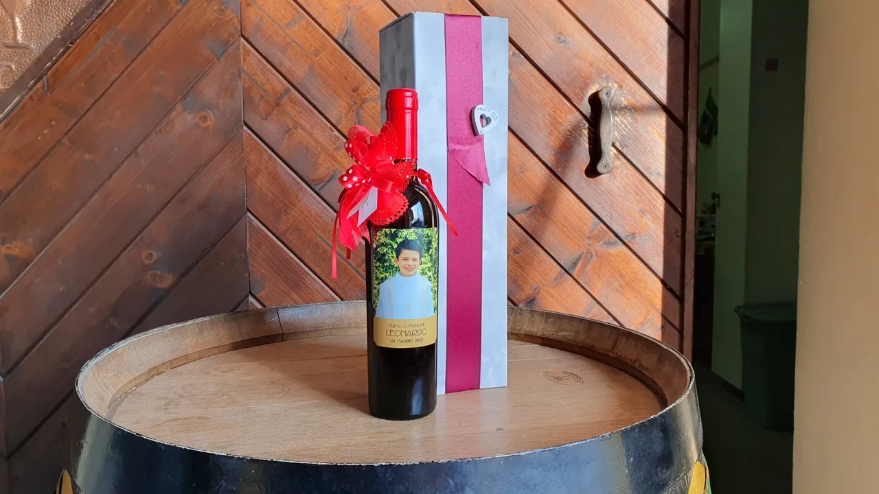Bottiglia formato speciale per comunione con etichetta personalizzata e fiocco, affiancata da una scatola usata per un matrimonio, il tutto sopra a una botte